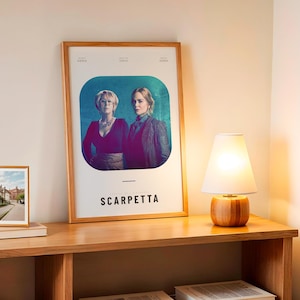 May include: A framed poster with the title "Scarpetta" featuring two women in a teal and purple background. A small framed photo and a wooden lamp with a white shade sit on a wooden table.