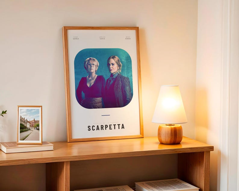 May include: A framed poster with the title "Scarpetta" featuring two women in a teal and purple background. A small framed photo and a wooden lamp with a white shade sit on a wooden table.