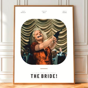 May include: A framed movie poster for "The Bride!" features a woman with dark makeup holding a gun. The poster includes the names Jessie Buckley, Christian Bale, and Annette Bening. The title "THE BRIDE!" is displayed in bold, capital letters.