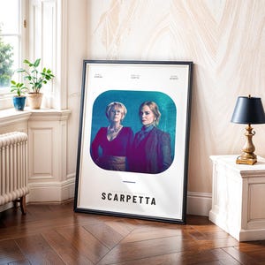 May include: A framed poster featuring two women against a teal background. The poster has the title "SCARPETTA" in large, bold letters. The poster is leaning against a wall in a room with a window and a small table with a lamp.