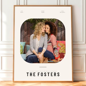 May include: A framed poster featuring two women embracing, set against a blurred outdoor backdrop. The poster includes the names of the actors: Teri Polo, Sherri Saum, and Maia Mitchell. The title "The Fosters" is displayed at the bottom.