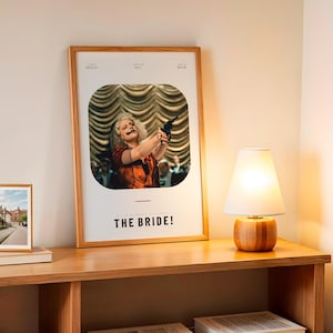 May include: A framed poster with the title "THE BRIDE!" features a woman holding a gun. A small framed photo and a wooden lamp with a white shade sit on a wooden table. The lamp has a wooden base.