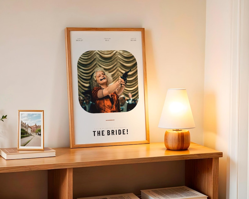 May include: A framed poster with the title "THE BRIDE!" features a woman holding a gun. A small framed photo and a wooden lamp with a white shade sit on a wooden table. The lamp has a wooden base.