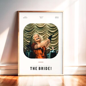 May include: Framed movie poster for "The Bride!" featuring a woman in an orange dress holding a gun. The poster has a white background with the movie title and actor names at the top and bottom. The frame is a light brown color.