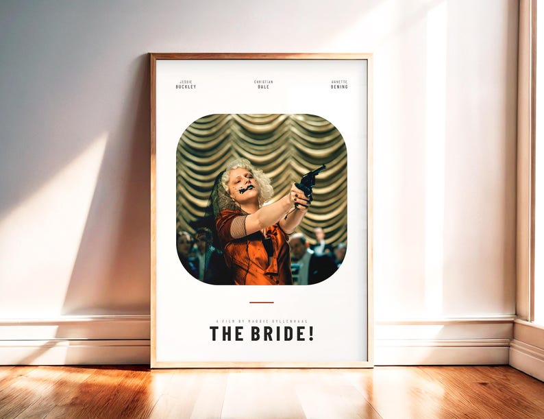 May include: Framed movie poster for "The Bride!" featuring a woman in an orange dress holding a gun. The poster has a white background with the movie title and actor names at the top and bottom. The frame is a light brown color.