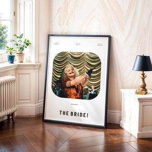May include: A framed movie poster for "THE BRIDE!" features a woman holding a gun. The poster has a black frame and is leaning against a wall. The room has a window, plants, and a small table with a lamp.