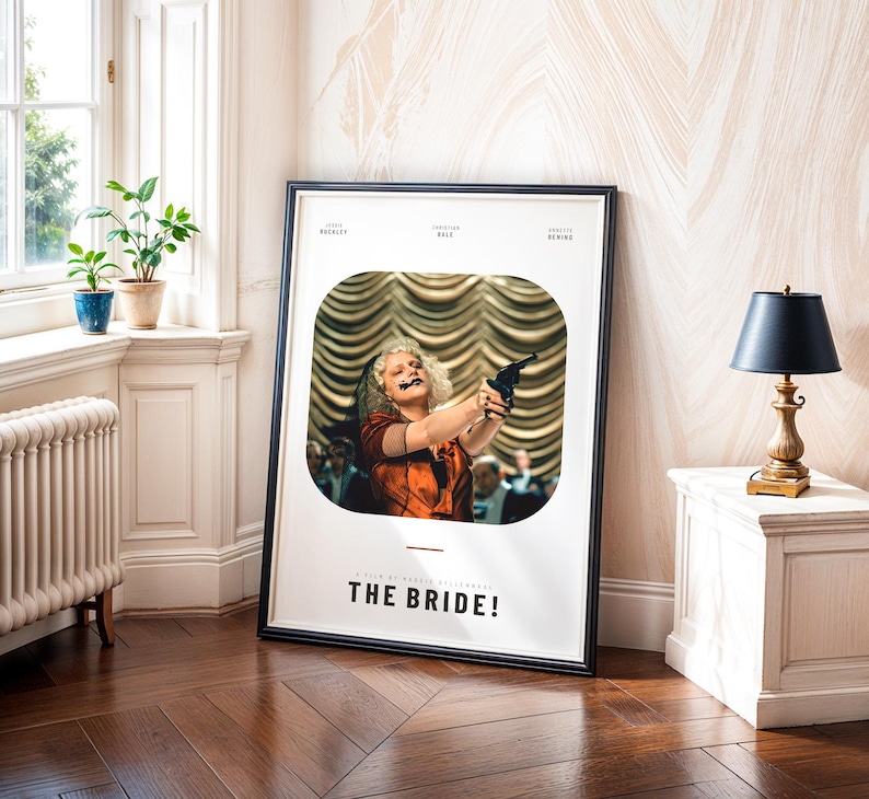 May include: A framed movie poster for "THE BRIDE!" features a woman holding a gun. The poster has a black frame and is leaning against a wall. The room has a window, plants, and a small table with a lamp.