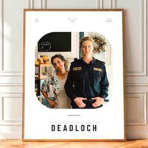 May include: A framed poster featuring two women. One woman is in a police uniform, and the other is wearing a floral shirt. The poster has the title "DEADLOCH" at the bottom and names at the top. The poster is in a wooden frame.