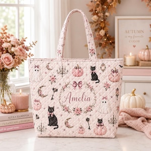 Könnte beinhalten: Eine gesteppte Tragetasche in hellem Rosa mit einem wiederkehrenden Muster aus schwarzen Katzen, Geistern, Kürbissen, Kronleuchtern und Schlüsseln. In der Mitte steht der Name Amelia in einem Blumenkranz mit Schleife. Die Tasche hat zwei Henkel.
