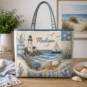 May include: A quilted tote bag featuring a coastal scene with a lighthouse, a sailboat, and seashells in shades of blue and cream. The name Madison is embroidered in blue script. The bag has two matching fabric handles and a rectangular shape.