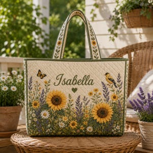 May include: A quilted tote bag featuring a cream-colored background with a printed garden scene of sunflowers, lavender, daisies, a butterfly, and a bird. The name Isabella is embroidered in green script above a small green heart.