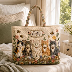 Op de afbeelding: Een gewatteerde tas met een geborduurde scène van vijf katten die achter een houten hek zitten, omringd door kleurrijke bloemen en vlinders. De naam Emily is geborduurd in een centraal decoratief kader. De hengsels hebben bloemmotieven.