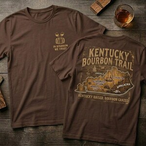 Puede incluir: Dos camisetas marrones con dise&ntilde;os tem&aacute;ticos de bourbon. Una camiseta presenta el texto "IN BOURBON WE TRUST" y un gr&aacute;fico de barril. La otra muestra un mapa del Kentucky Bourbon Trail con el texto "KENTUCKY BOURBON TRAIL".