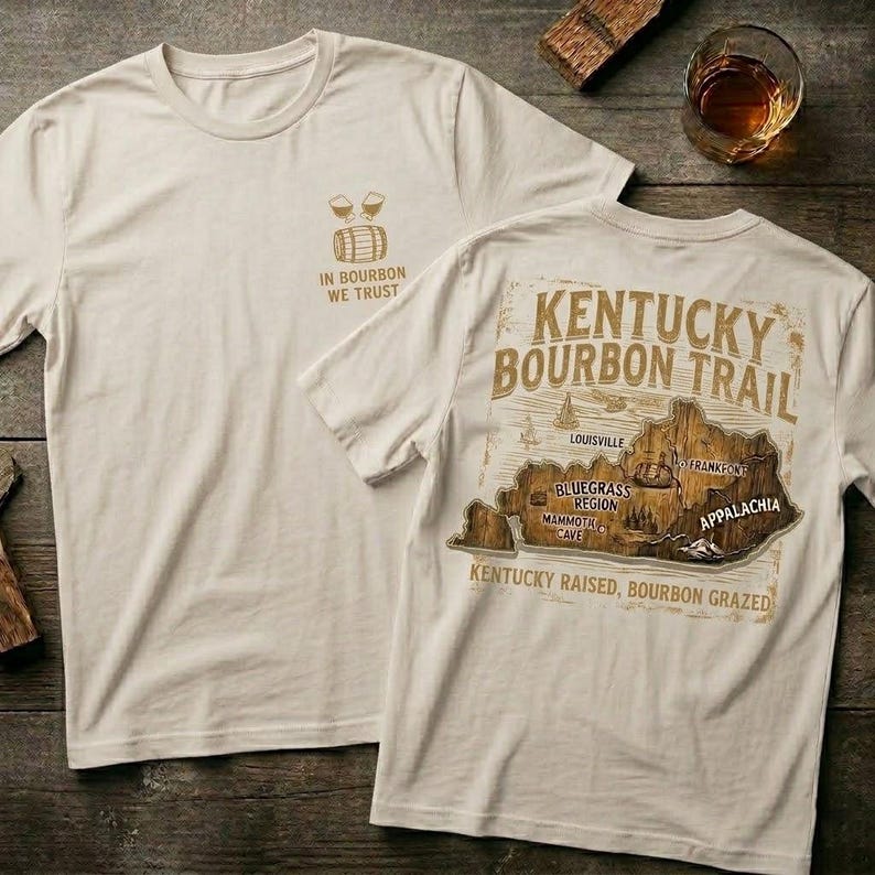 Puede incluir: Dos camisetas color crema. Una camiseta presenta un peque&ntilde;o gr&aacute;fico con el texto "IN BOURBON WE TRUST". La otra camiseta tiene un gr&aacute;fico de mapa con el texto "KENTUCKY BOURBON TRAIL" y "KENTUCKY RAISED, BOURBON GRAZED".