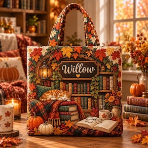 Op de afbeelding: Een draagtas met een gewatteerd herfsttafereel met een slapende oranje cyperse kat, een boekenplank, pompoenen en een lantaarn. De naam Willow staat op een centraal houten bord. Het ontwerp bevat levendige herfstbladeren, een boek en een bril.