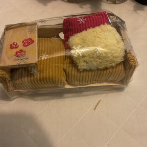 Può includere: Un set di divano in miniatura in velluto a coste con un tavolino abbinato e due cuscini, il tutto confezionato in plastica trasparente. Il divano e il tavolino sono marrone chiaro e i cuscini sono rosa e gialli. Il tavolino ha un motivo floreale.