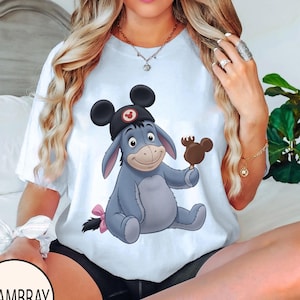 May include: White t-shirt featuring a cartoon illustration of Eeyore wearing a Mickey Mouse hat and holding a Mickey Mouse-shaped ice cream bar. Eeyore is light blue with a pink bow on his tail. The shirt has a round neckline.