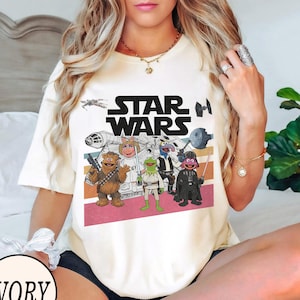 May include: Ivory t-shirt with the words "STAR WARS" and a graphic of Star Wars characters and Muppets. The graphic includes Chewbacca, Miss Piggy, Kermit the Frog, Gonzo, and Darth Vader. The design has retro horizontal stripes.
