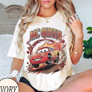 Puede incluir: Una camiseta de manga corta color marfil con un estampado gr&aacute;fico del personaje de coche de carreras rojo Lightning McQueen y el texto MC QUEEN encima. El dise&ntilde;o incluye llamas, llaves inglesas y el n&uacute;mero 95 en el coche sobre un fondo polvoriento.