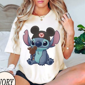 K&ouml;nnte beinhalten: Elfenbeinfarbenes T-Shirt mit einer Cartoon-Figur Stitch, die einen Mickey Mouse-Hut tr&auml;gt und einen Schokoladeneisriegel h&auml;lt. Das Design enth&auml;lt das Wort "IVORY" in einem kreisf&ouml;rmigen Schriftzug unten links.