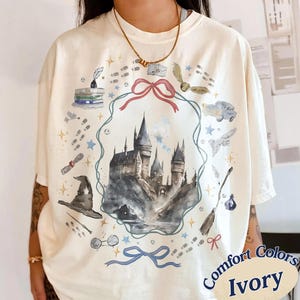 May include: An ivory t-shirt featuring a watercolor-style graphic of a large castle surrounded by a decorative frame with a pink bow at the top and blue bow at the bottom. The design includes illustrations of a wizard hat, books, a broom, and stars.