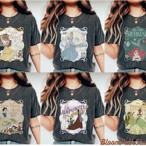 Op de afbeelding: Een collectie van zes antracietgrijze T-shirts met korte mouwen, elk voorzien van een andere vintage-stijl grafische print van een sprookjesfiguur, waaronder Belle en het Beest, Assepoester, De Kleine Zeemeermin, Sneeuwwitje, Rapunzel en Tiana.