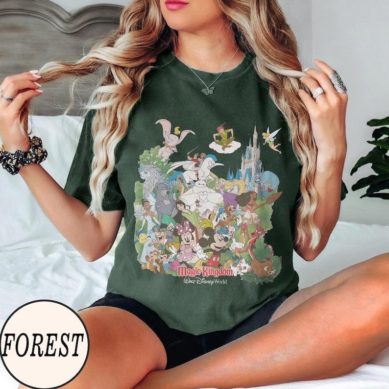 Peut inclure: T-shirt vert fonc&eacute; avec un graphisme color&eacute; de personnages Disney, dont Mickey Mouse, Dumbo et le ch&acirc;teau de Cendrillon. Le texte "Magic Kingdom Walt Disney World" est imprim&eacute; en dessous. Le t-shirt a des manches courtes.