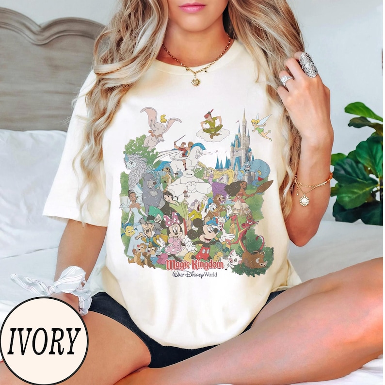 Peut inclure: T-shirt ivoire avec un graphisme color&eacute; de personnages Disney, dont Mickey Mouse, Minnie Mouse et Peter Pan. Le graphisme comprend le texte "Magic Kingdom" et "Walt Disney World".