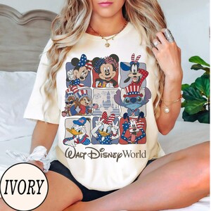 May include: Ivory-colored t-shirt featuring a grid of Disney characters in patriotic attire. Mickey Mouse, Minnie Mouse, and Donald Duck are among the characters. The text "Walt Disney World" is printed below the grid.