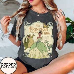Op de afbeelding: Een antracietgrijs T-shirt met korte mouwen en een grafische print van The Princess and the Frog. Het ontwerp bevat Tiana, de alligator Louis en een kikker, met de tekst The Princess and the Frog en The Real Magic Is Believing In You.