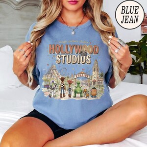 May include: A light blue crewneck sweatshirt featuring a graphic print of Hollywood Studios with Toy Story characters including Buzz Lightyear, Woody, Jessie, Rex, Slinky Dog, and Mr. Potato Head. The text reads magic picture studio HOLLYWOOD STUDIOS.