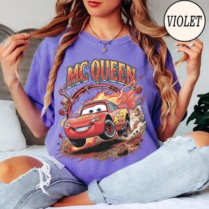 Puede incluir: Una camiseta violeta con un gr&aacute;fico de un coche de carreras rojo con el n&uacute;mero 95 en el lateral, rodeado de llamas, engranajes y llaves inglesas. El texto MC QUEEN est&aacute; impreso en una fuente negrita y arqueada sobre el coche.