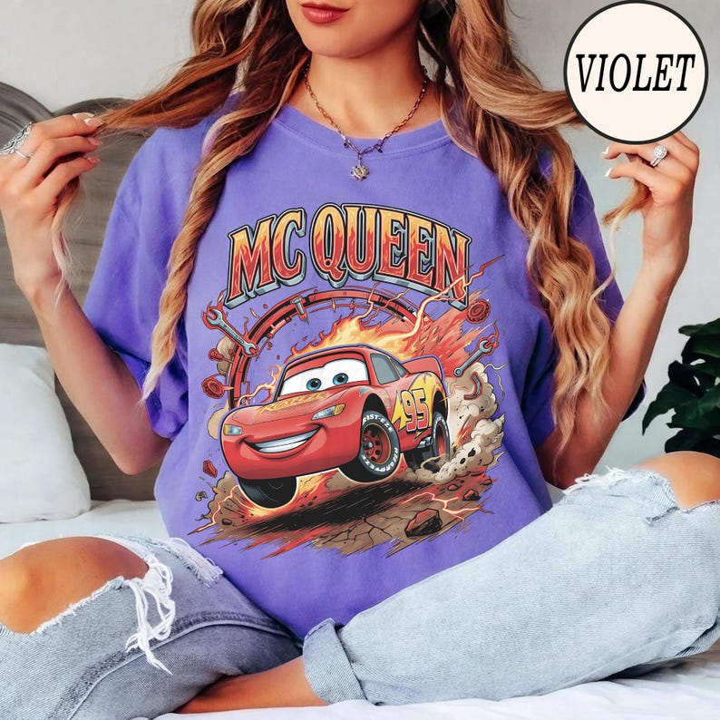 Puede incluir: Una camiseta violeta con un gr&aacute;fico de un coche de carreras rojo con el n&uacute;mero 95 en el lateral, rodeado de llamas, engranajes y llaves inglesas. El texto MC QUEEN est&aacute; impreso en una fuente negrita y arqueada sobre el coche.