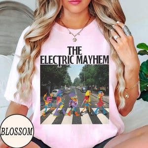 Pode incluir: T-shirt rosa claro com a frase "The Electric Mayhem". O gr&aacute;fico mostra personagens dos Muppets atravessando uma rua, com uma estrada e &aacute;rvores ao fundo. A camiseta tem mangas curtas.