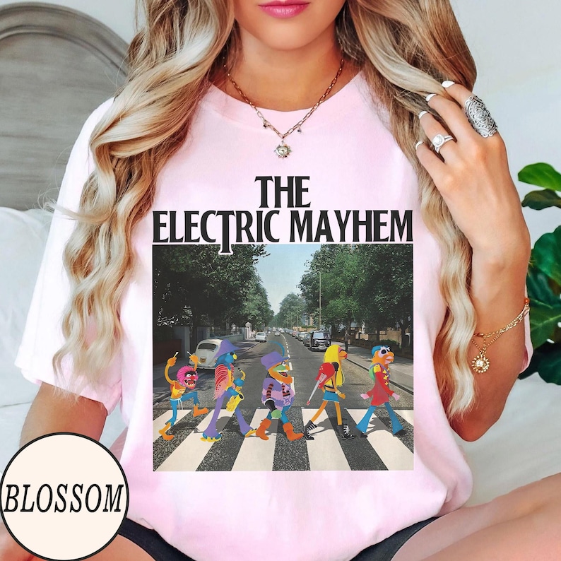 Pode incluir: T-shirt rosa claro com a frase "The Electric Mayhem". O gr&aacute;fico mostra personagens dos Muppets atravessando uma rua, com uma estrada e &aacute;rvores ao fundo. A camiseta tem mangas curtas.