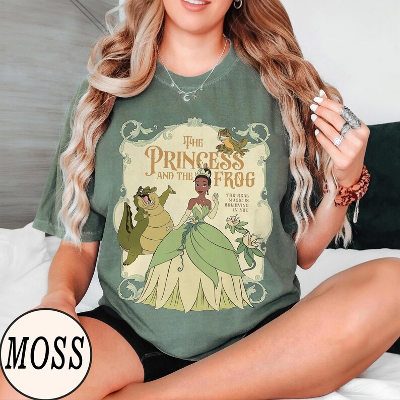 Op de afbeelding: Een mosgroen t-shirt met korte mouwen en een grafische print van The Princess and the Frog. Het ontwerp toont Tiana, de alligator Louis en een kikker in een sierlijk kader, met de tekst The Princess and the Frog en The Real Magic Is Believing In You.