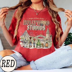 May include: A red short-sleeve t-shirt featuring a graphic print with the text magic picture studio HOLLYWOOD STUDIOS and a film clapperboard icon. The design includes illustrations of various animated characters standing before a theme park entrance.