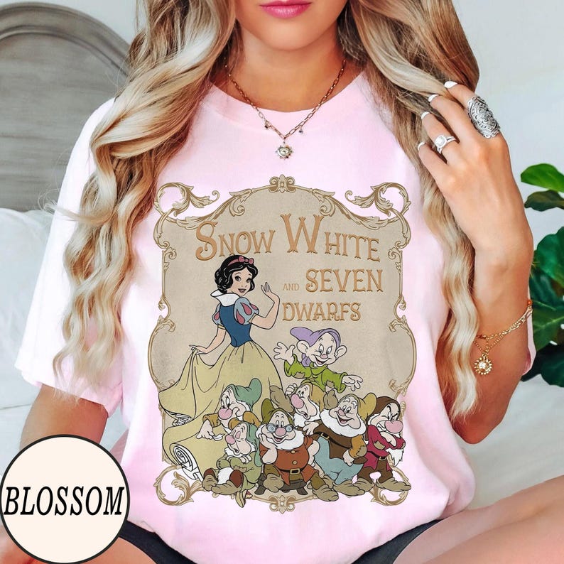 Op de afbeelding: Een lichtroze t-shirt met een grafische print van Sneeuwwitje en de zeven dwergen in een sierlijk, beige decoratief kader. De tekst "Snow White and Seven Dwarfs" staat in een bruin, gestileerd lettertype boven de personages gedrukt.