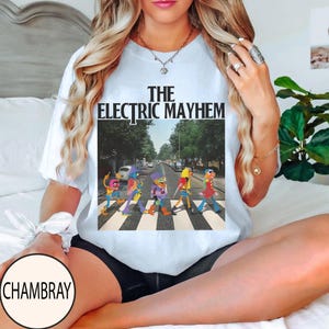 Pode incluir: Camiseta branca com a frase "THE ELECTRIC MAYHEM" acima de um gr&aacute;fico colorido de personagens dos Muppets atravessando uma rua. A imagem &eacute; uma par&oacute;dia da capa do &aacute;lbum Abbey Road dos Beatles.