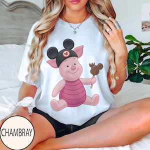 May include: White t-shirt featuring a cartoon pig wearing a black Mickey Mouse hat and holding a chocolate-covered Mickey Mouse-shaped treat. The pig is pink with a striped pink shirt. The shirt has a round neck.