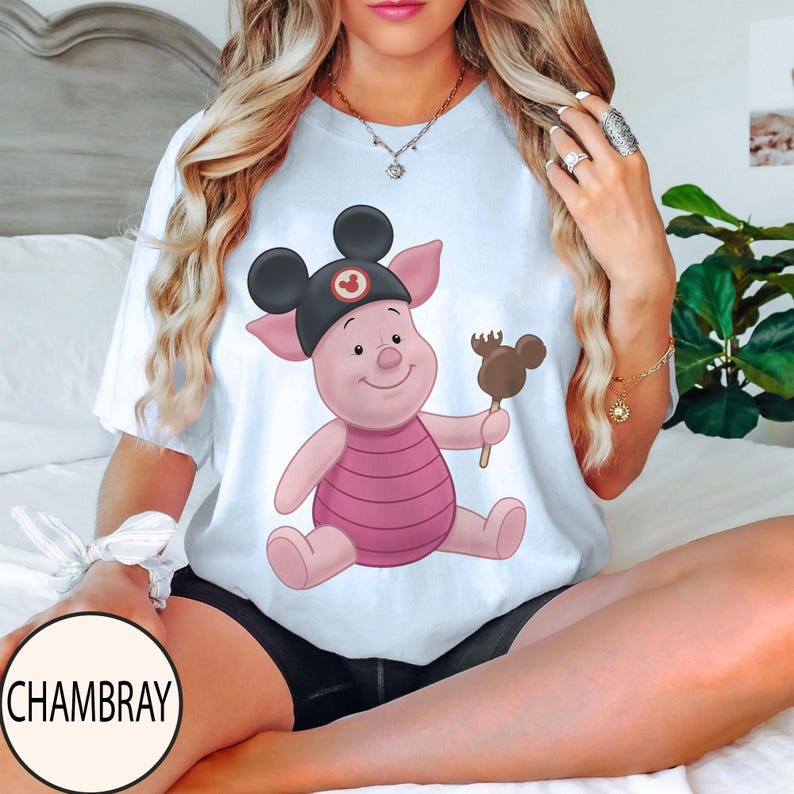 May include: White t-shirt featuring a cartoon pig wearing a black Mickey Mouse hat and holding a chocolate-covered Mickey Mouse-shaped treat. The pig is pink with a striped pink shirt. The shirt has a round neck.