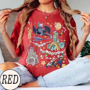 May include: Red t-shirt with a colorful graphic design featuring a dragon, castle, and video game characters. The words "EPIC UNIVERSE" are displayed in a circular pattern. The shirt is made of a soft material.