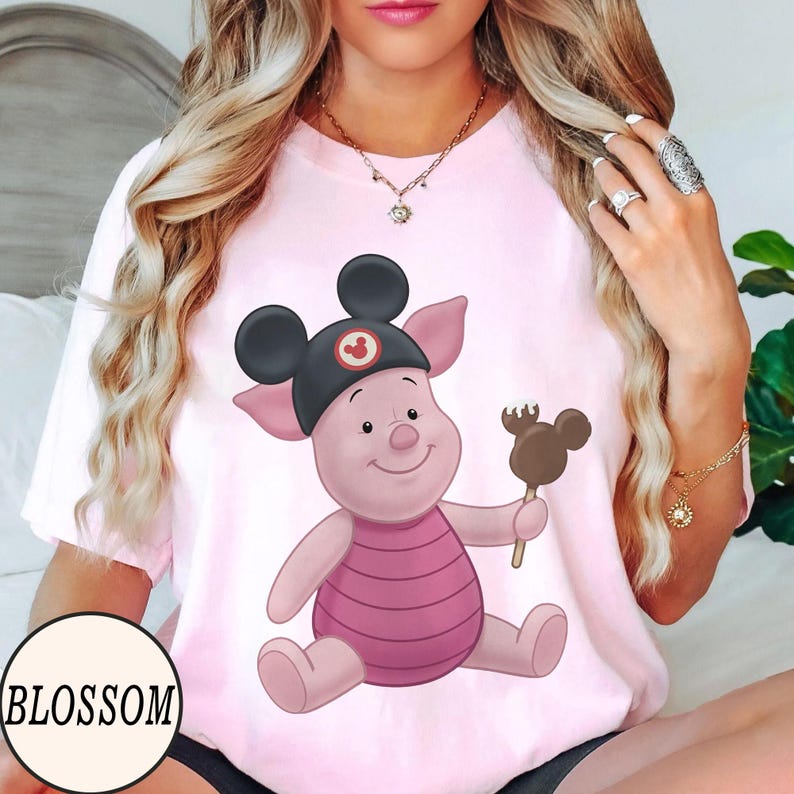 May include: Pink t-shirt featuring a cartoon pig wearing a Mickey Mouse hat and holding a chocolate-covered ice cream bar. The word "BLOSSOM" is printed in a black font at the bottom left of the shirt.