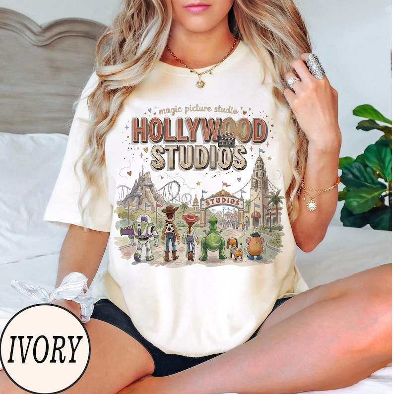 May include: An ivory short-sleeve t-shirt featuring a graphic print with the text "magic picture studio HOLLYWOOD STUDIOS" above an illustration of various animated characters standing before a theme park entrance with a roller coaster.