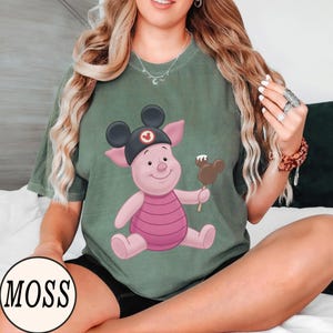 May include: Sage green t-shirt featuring a cartoon Piglet character wearing a Mickey Mouse hat and holding a chocolate-covered ice cream bar. The shirt has a round neckline and short sleeves. The word "MOSS" is printed in a circle at the bottom left.