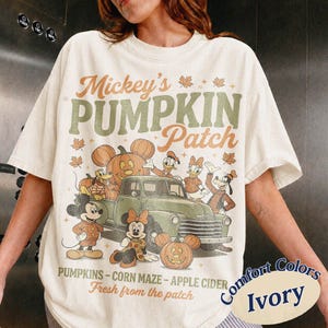 Könnte beinhalten: Ein elfenbeinfarbenes T-Shirt mit einem Grafikdruck, der den Text Mickey's PUMPKIN Patch, PUMPKINS - CORN MAZE - APPLE CIDER und Fresh from the patch enthält. Das Motiv zeigt Mickey Maus, Minnie Maus, Donald Duck, Daisy Duck, Goofy und Pluto.