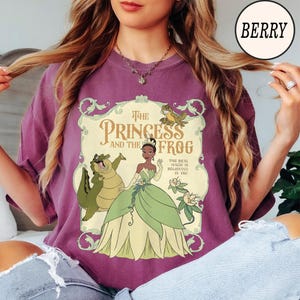 Op de afbeelding: Een bessenkleurig t-shirt met een grafische print van Tiana uit The Princess and the Frog, samen met Louis de alligator en Ray de vuurvlieg. De tekst luidt The Princess and the Frog en The Real Magic is Believing in You.