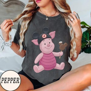 May include: Dark gray t-shirt featuring a cartoon Piglet character wearing a Mickey Mouse hat and holding a Mickey Mouse-shaped ice cream bar. The character is pink with a pink and white striped shirt. The word "PEPPER" is in a white circle at the bottom.