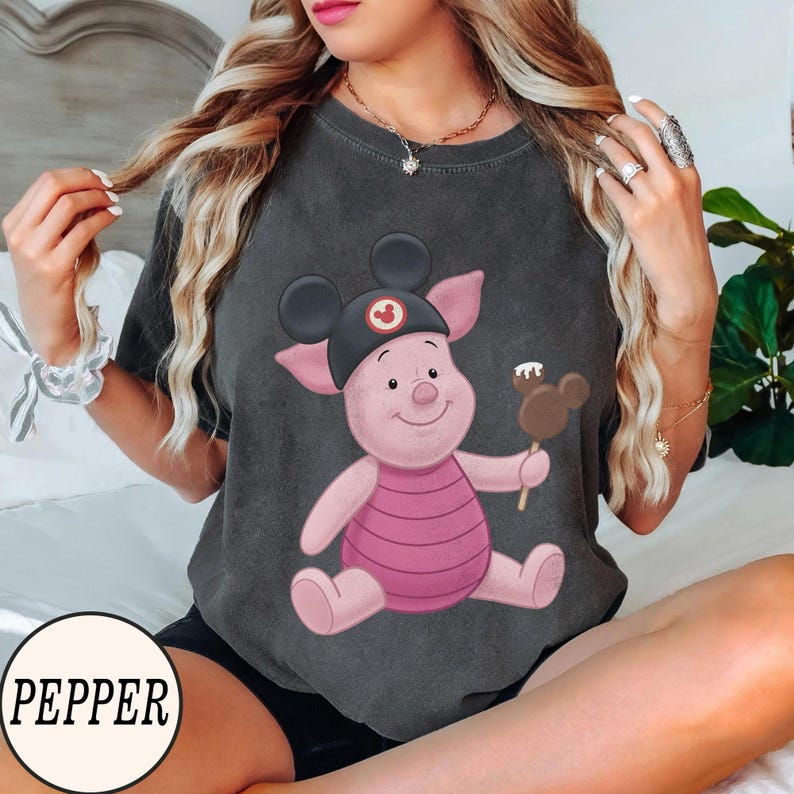 May include: Dark gray t-shirt featuring a cartoon Piglet character wearing a Mickey Mouse hat and holding a Mickey Mouse-shaped ice cream bar. The character is pink with a pink and white striped shirt. The word "PEPPER" is in a white circle at the bottom.
