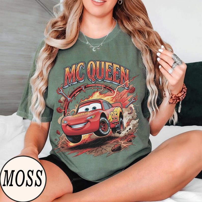 Puede incluir: Una camiseta de manga corta color verde musgo con un gr&aacute;fico del personaje de coche de carreras rojo Lightning McQueen y el texto MC QUEEN encima. El dise&ntilde;o incluye llamas, llaves inglesas y el n&uacute;mero 95 en el coche, sobre un fondo polvoriento.